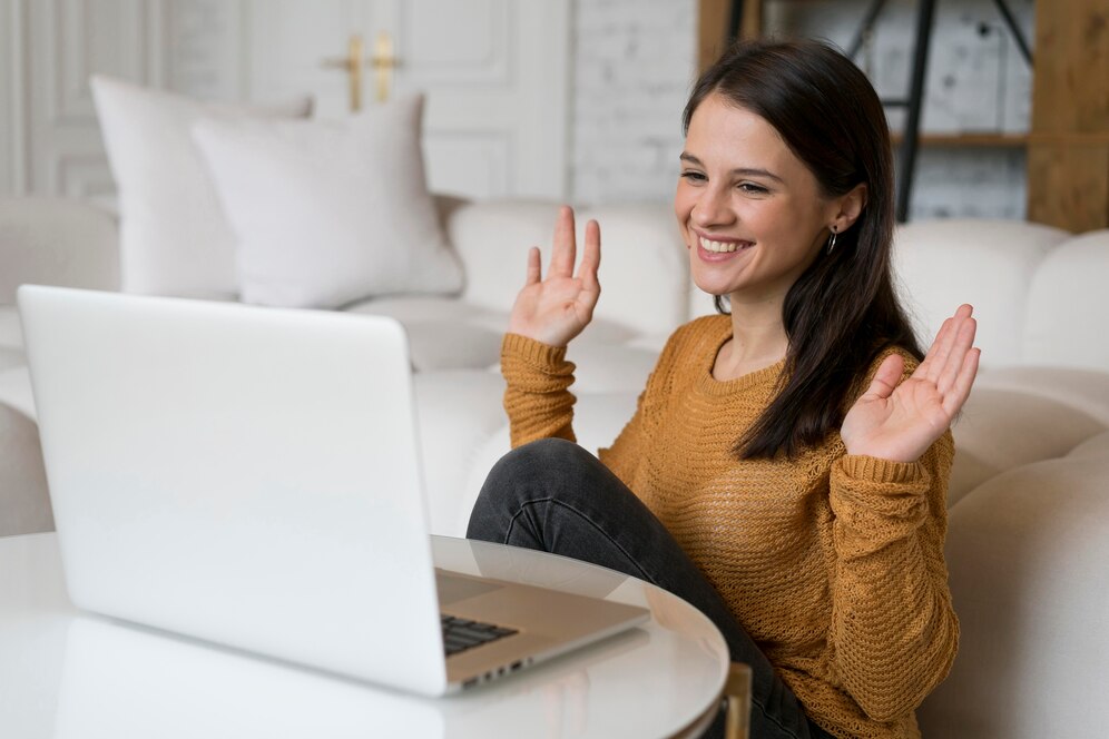 Woman sitting on a sofa with a white laptop on a round table, smiling and waving at the screen.
