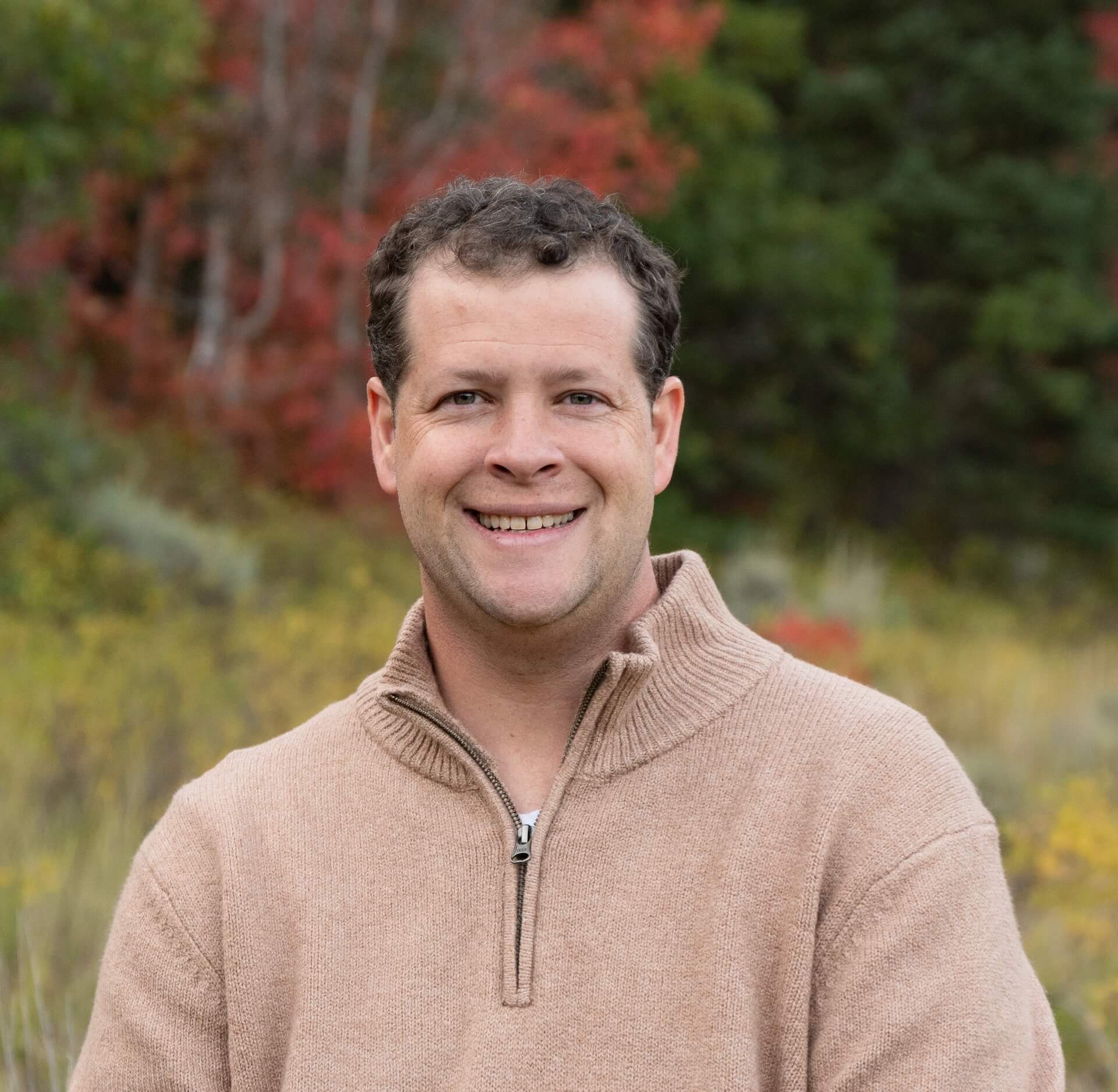 Smiling man wearing a beige zip-neck sweater outdoors with fall foliage in the blurred background.