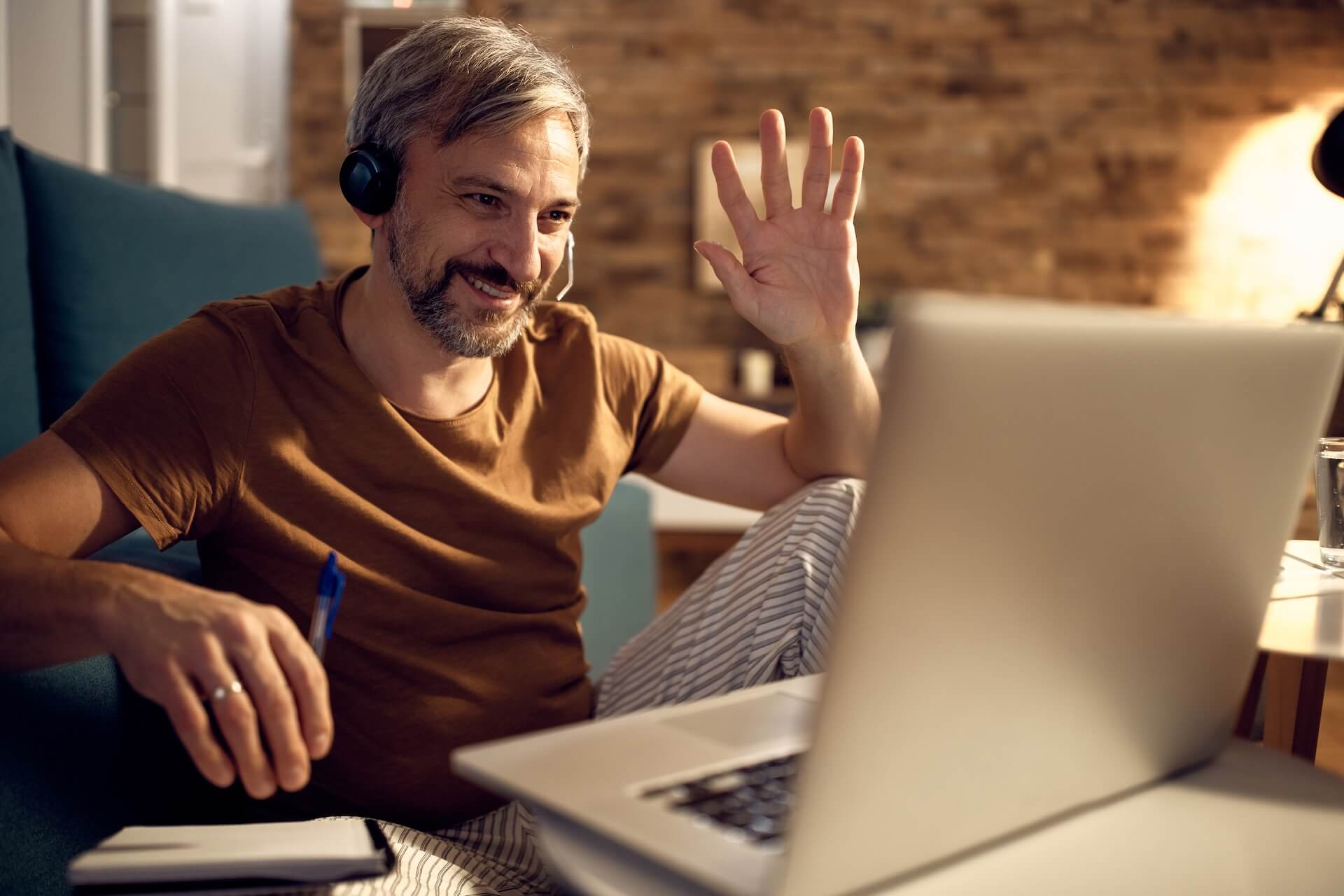 Smiling man wearing a headset waves at a laptop in a home office setting.