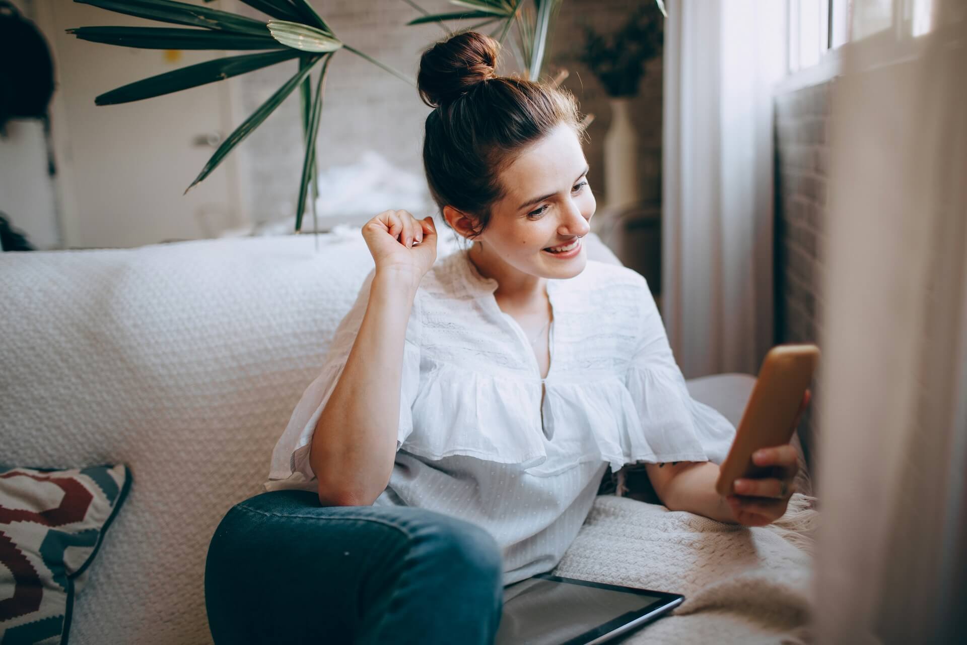 Woman sitting on a sofa, looking at her smartphone with a smile, in a bright living room.