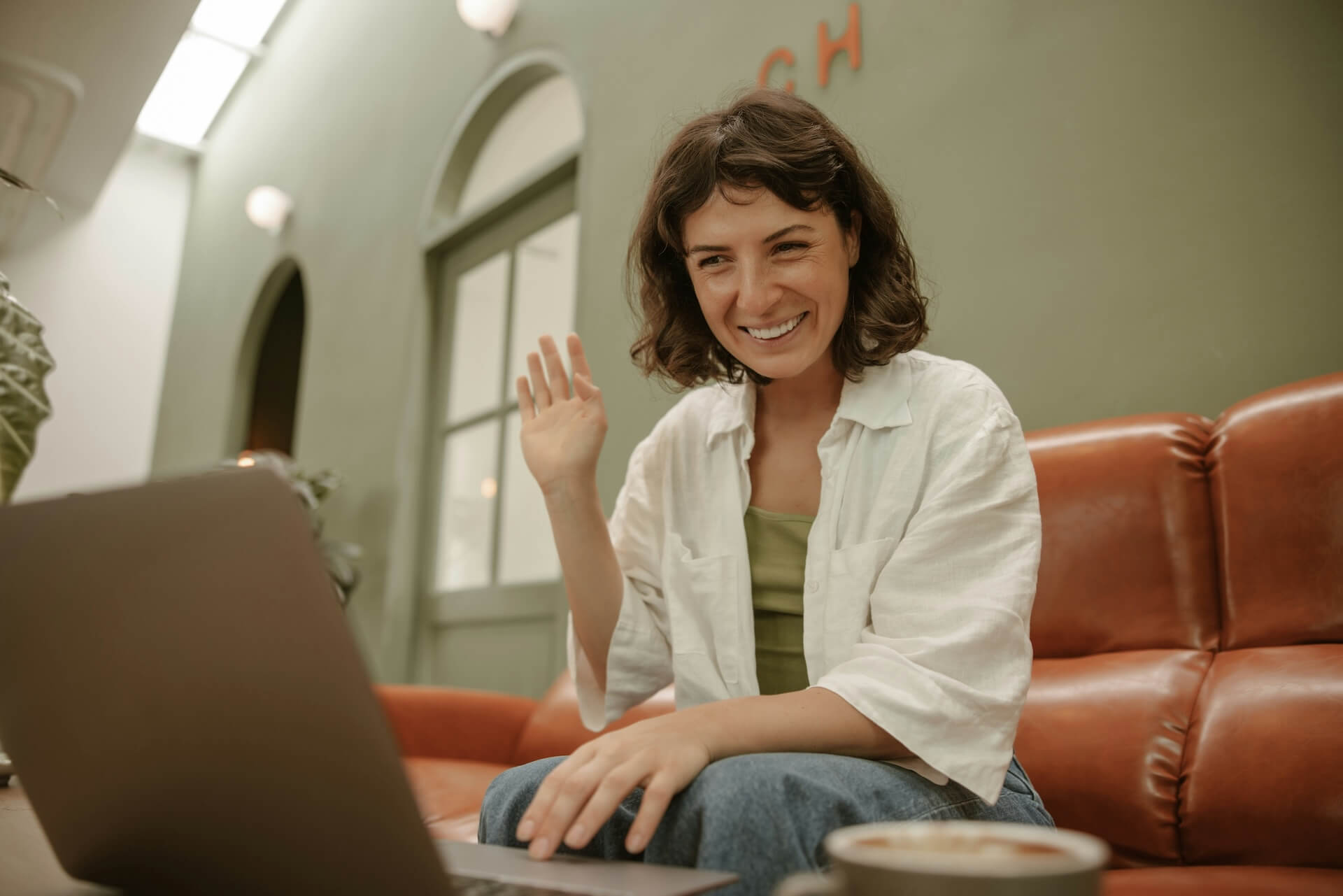 Smiling woman in a white shirt waves at a laptop on a coffee table in a cozy lounge.