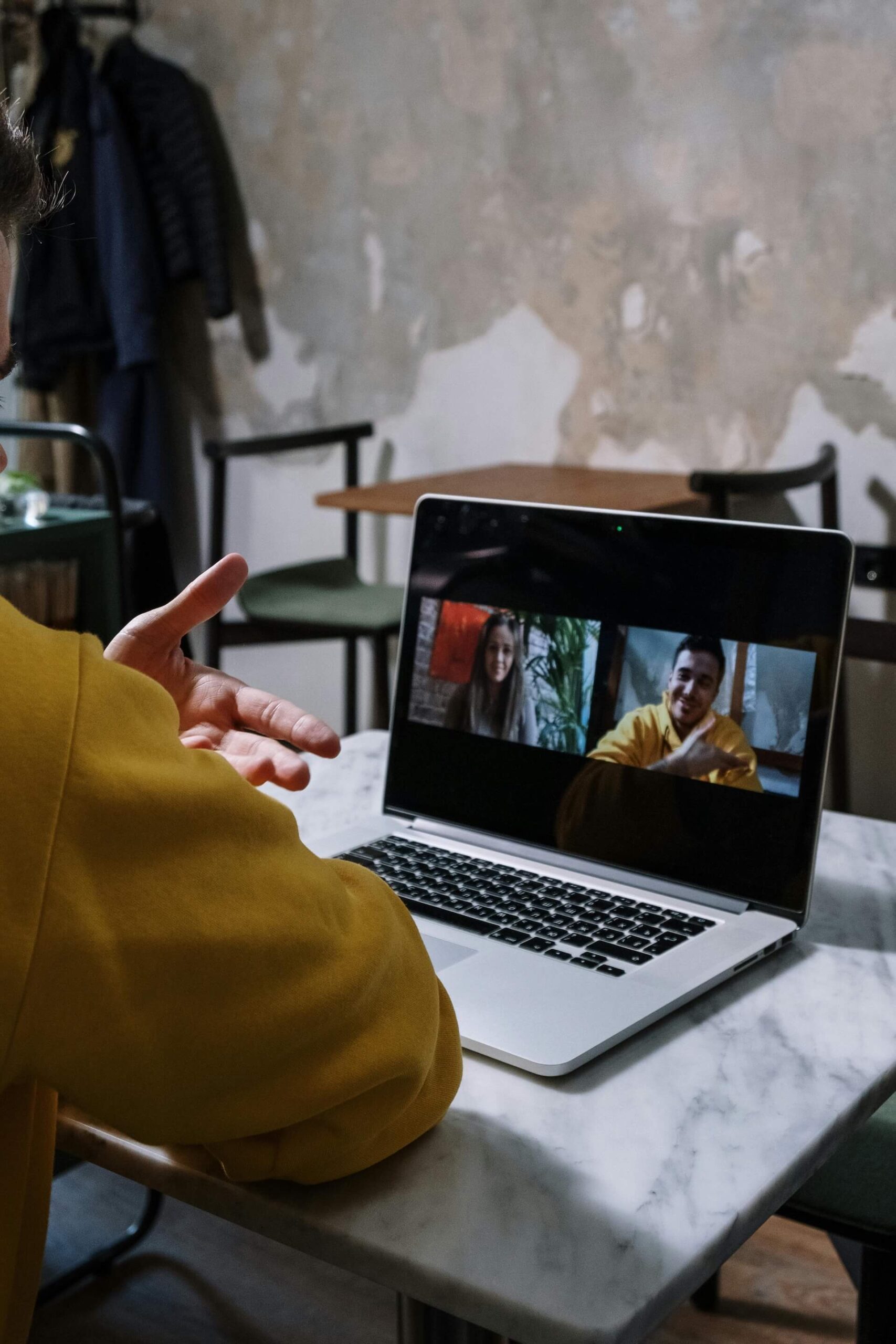 Person in a yellow hoodie sits at a table, holding out hand toward a laptop showing a video call with four people.