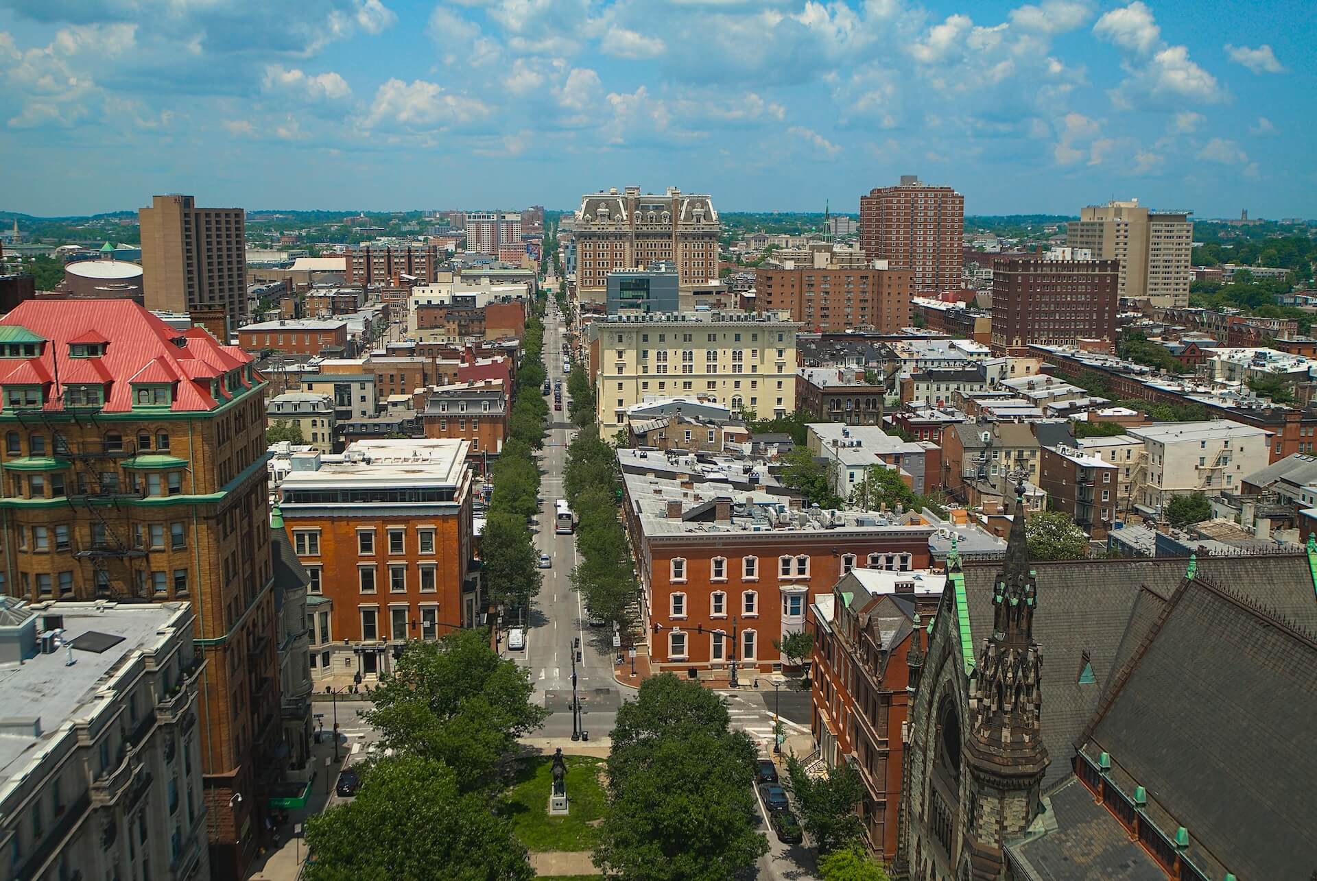 Aerial cityscape with a tree-lined avenue and varied buildings, extending toward a distant skyline under a blue sky and fluffy clouds.