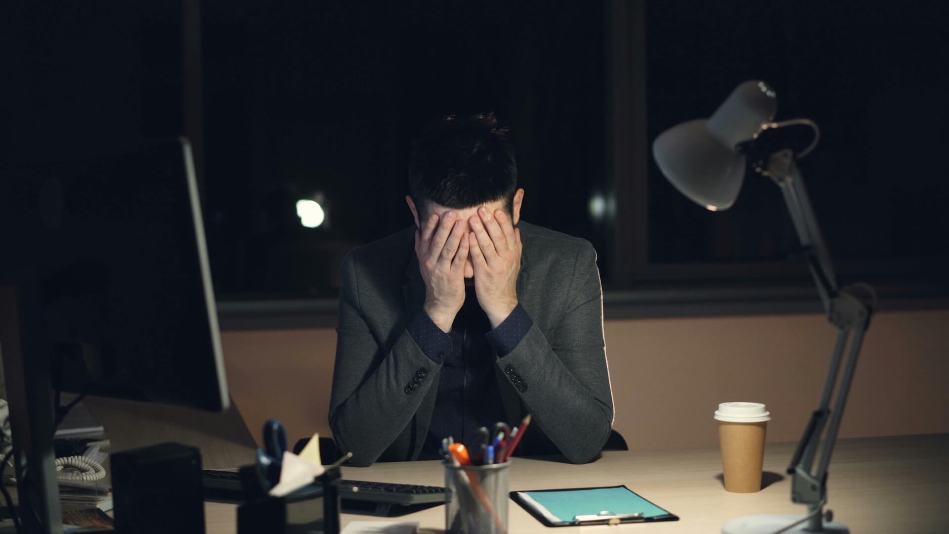 Man in a blazer sits at a dimly lit desk at night, covering his face with both hands in apparent frustration or fatigue, with a coffee cup and office equipment nearby.