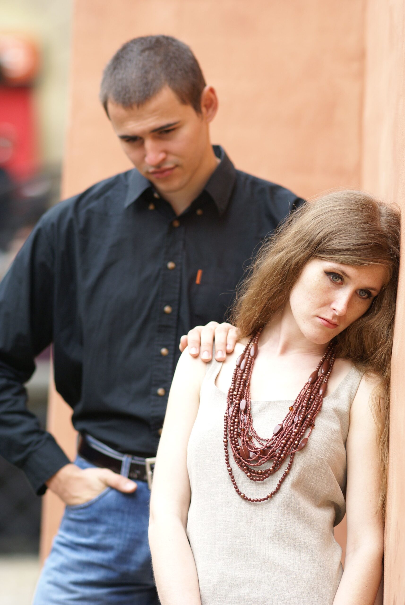 Man in a black button-down shirt stands behind a woman with long brown hair, who wears a beaded brown necklace and beige sleeveless dress, leaning against a peach wall.