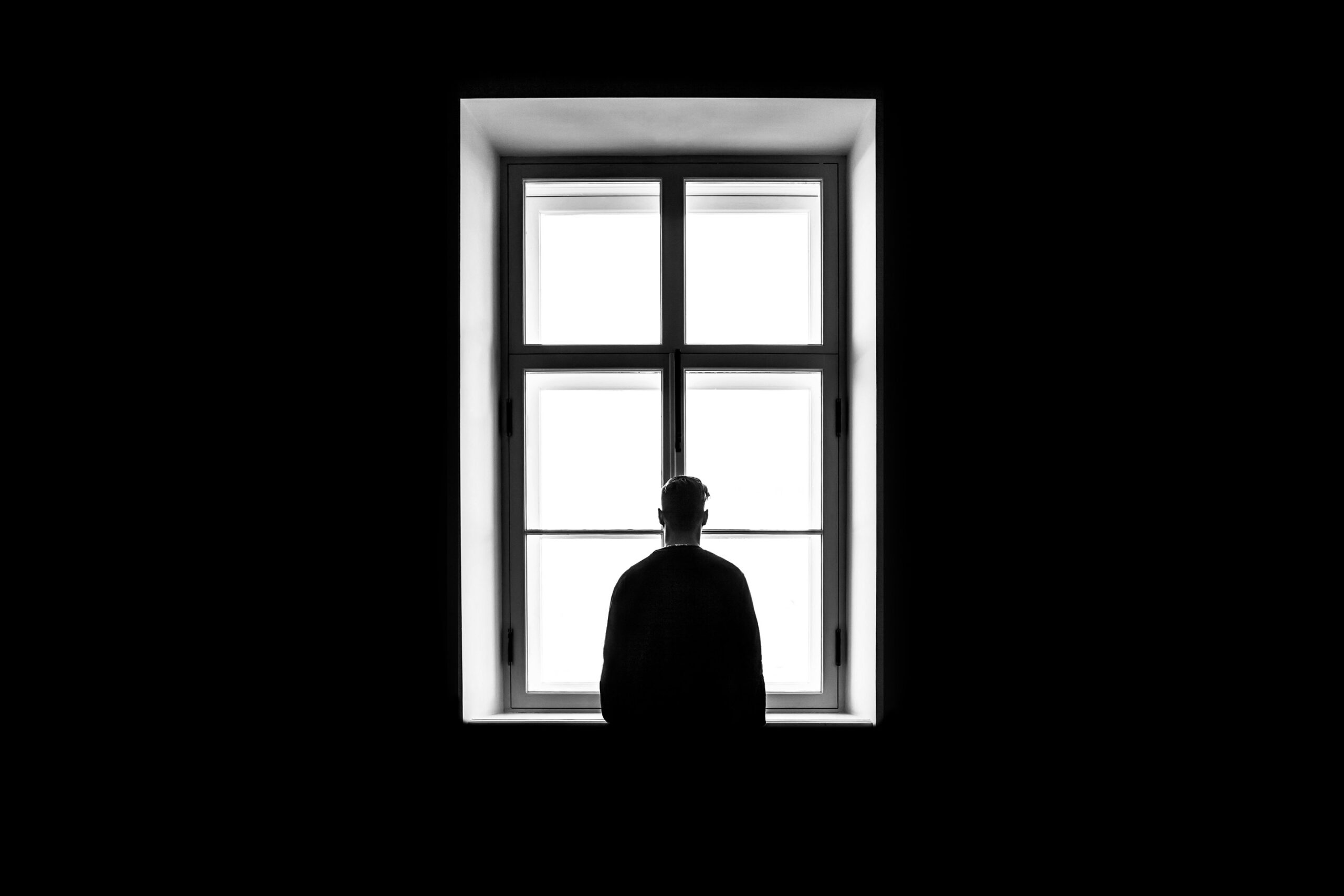 Silhouette of a person standing with back to the camera in front of a bright four-pane window in a dark room.