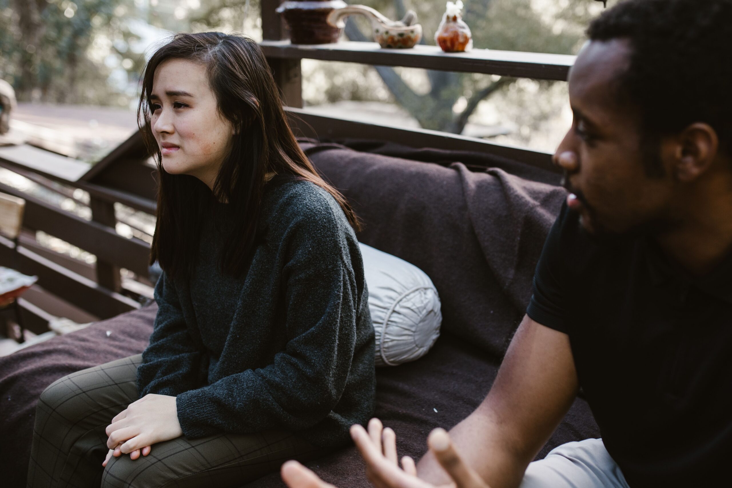 Two people sitting on a wooden porch swing outdoors, with the woman in a gray sweater looking away and the man partially visible on the right.